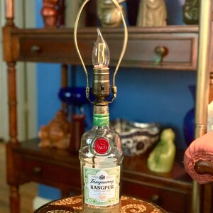 May include: A clear glass bottle lamp with a gold metal lampshade. The bottle is labeled "Tanqueray Rangpur Distilled Gin". The lamp is sitting on a small, round table with a mosaic tile top.