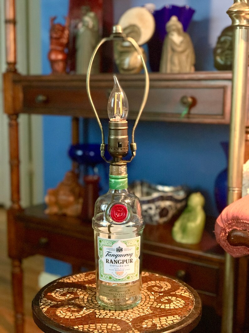 May include: A clear glass bottle lamp with a gold metal lampshade. The bottle is labeled "Tanqueray Rangpur Distilled Gin". The lamp is sitting on a small, round table with a mosaic tile top.