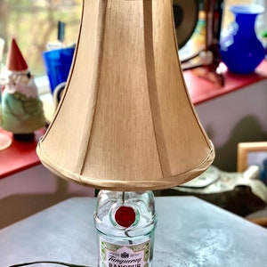 May include: A table lamp made from a repurposed Tanqueray Rangpur gin bottle with a beige fabric lampshade. The bottle has a red wax seal on the top.