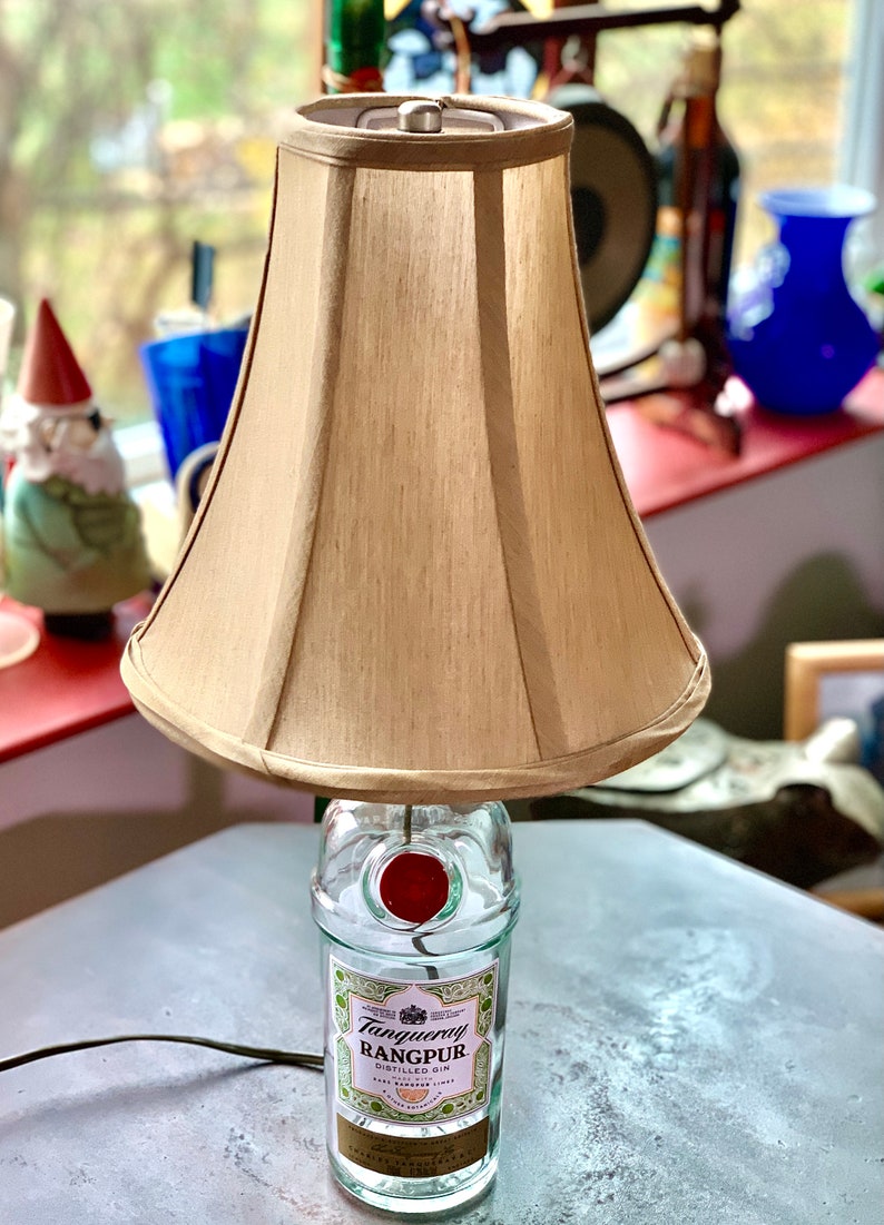 May include: A table lamp made from a repurposed Tanqueray Rangpur gin bottle with a beige fabric lampshade. The bottle has a red wax seal on the top.