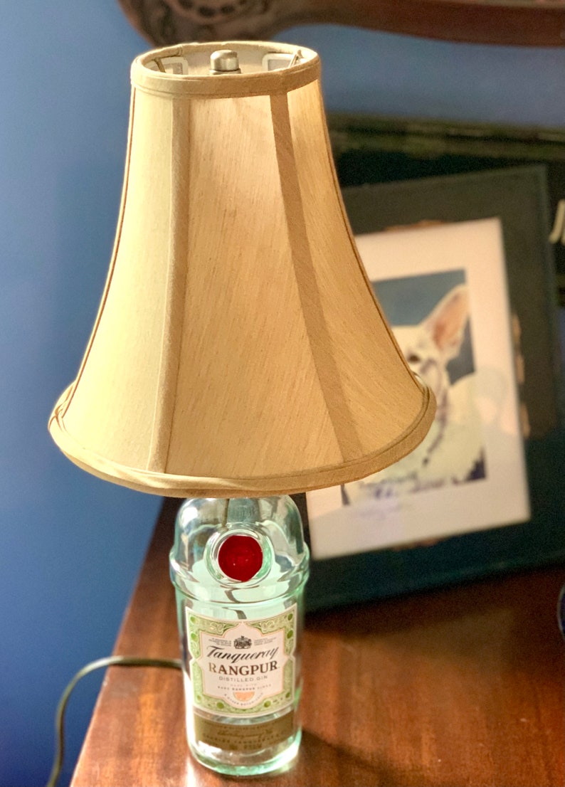 May include: A table lamp made from a clear glass bottle with a red cap. The bottle has a label that reads "Tanqueray Rangpur Distilled Gin". The lamp has a beige fabric lampshade.
