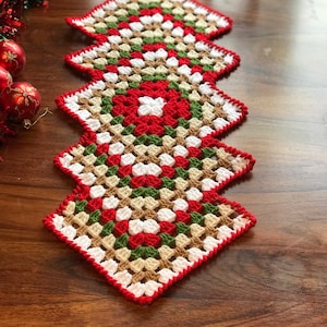 May include: A crocheted Christmas tree-shaped table runner. The runner features a granny square design with red, white, green, and tan squares. The edges are trimmed with a red border. Christmas ornaments and garland are in the background.