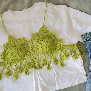 May include: A lime green crochet top with delicate straps and fringe detailing. The top is displayed on a white t-shirt, with a pair of blue denim shorts visible in the background. The crochet design features a scalloped edge.