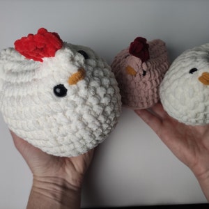 May include: Three crocheted chicken plushies. One is white with a red comb, one is white with a red comb and a yellow beak, and one is pink with a maroon comb.