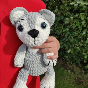 May include: A handmade plush toy wolf with gray and white yarn. The wolf has large, expressive eyes with green irises, a black nose, and black claws. The toy is held against a red shirt and green foliage.
