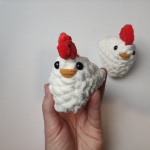 May include: Two white crocheted chickens with red combs and black eyes. The chickens are sitting on a white surface.