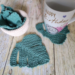 May include: Three teal green crocheted coasters with a white crocheted bowl filled with more teal green crocheted coasters. A white mug with the text "Happy Birthday" is sitting on one of the coasters.