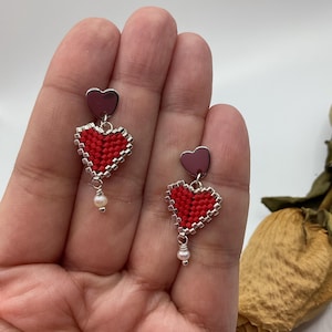 May include: A pair of heart-shaped earrings. Each earring features a small, red beaded heart with a silver border, topped with a small, dark pink heart. A small pearl dangles from the bottom of each earring. The earrings are displayed against a white background.