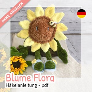 May include: A crocheted sunflower toy with a brown face, yellow petals, and green leaves. The toy is sitting on a white surface with a small crocheted bee on its head. The text "Blume Flora Häkelanleitung - pdf" is written below the toy.