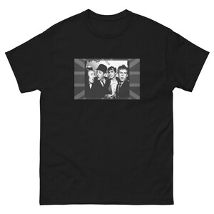 May include: Black and white photo of four men in a band, wearing suits and ties, standing in front of a Union Jack flag. The photo is printed on a black t-shirt.