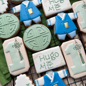 May include: An assortment of decorated cookies, including blue and white traditional Korean outfits, green circular designs, the number 1, and cookies with the name "Hugo" in green lettering. The cookies are arranged on a black wire rack.