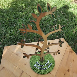 May include: A wooden tree cutout with green leaves and a green glitter base. The base has the text "Happy 22nd Birthday Alec" written on it.