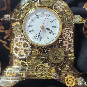 Könnte beinhalten: Eine transparente Harz-Uhr mit goldfarbenen Zahnrädern und einem weißen Zifferblatt mit schwarzen römischen Ziffern. Die Uhrzeiger sind schwarz und rot.