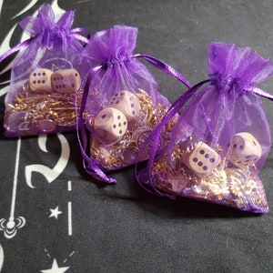 Op de afbeelding: Drie paarse organza zakjes gevuld met gouden kettingen en houten dobbelstenen met paarse stippen. De zakjes zijn gebonden met een paars lint.