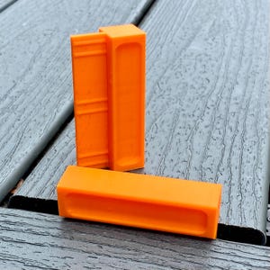 May include: Two orange plastic corner clamps for woodworking projects. The clamps are made of a durable plastic and are designed to hold pieces of wood together at a 90-degree angle.