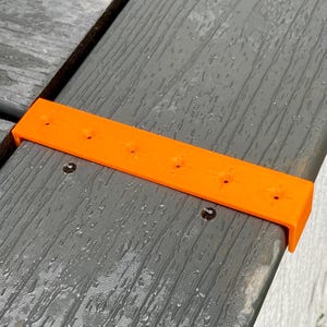 May include: Orange plastic deck board support bracket with four holes for screws. The bracket is attached to a gray composite deck board.