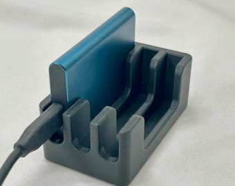 May include: A teal-colored external hard drive is docked in a black 3D-printed holder. A black USB cable is plugged into the hard drive. The holder has four slots and is designed to keep the hard drive upright.
