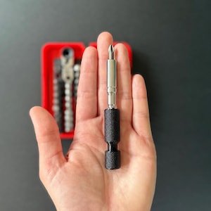 May include: A black and silver screwdriver bit held in a hand. The bit has a textured black handle and a silver shaft. A red case with other bits is in the background.