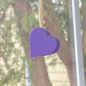 May include: A purple heart-shaped object hanging from a white string. The heart is made of a textured material and is hanging in front of a blurred background.