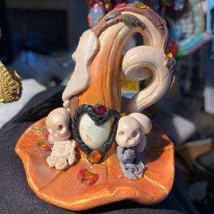 May include: A miniature witch's hat made of orange, brown, and white clay with a swirl design. The hat is decorated with small, colorful gems and two small, white skull figures.