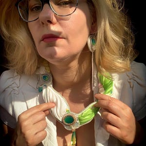 May include: A woman wearing a white blouse and a green and white necklace with a large green stone pendant. She is also wearing matching earrings with green stones. The necklace and earrings appear to be handmade.