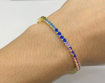 May include: A gold bracelet with a rainbow of colorful stones. The stones are set in a gold band and are arranged in a gradient from yellow to pink.