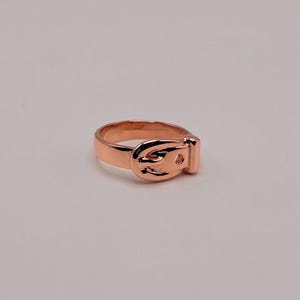 May include: A rose gold-toned metal ring designed to resemble a miniature belt. The band features a central buckle element with a prong and a small hole, mimicking the appearance of a functional belt wrapped around the finger in a smooth finish.