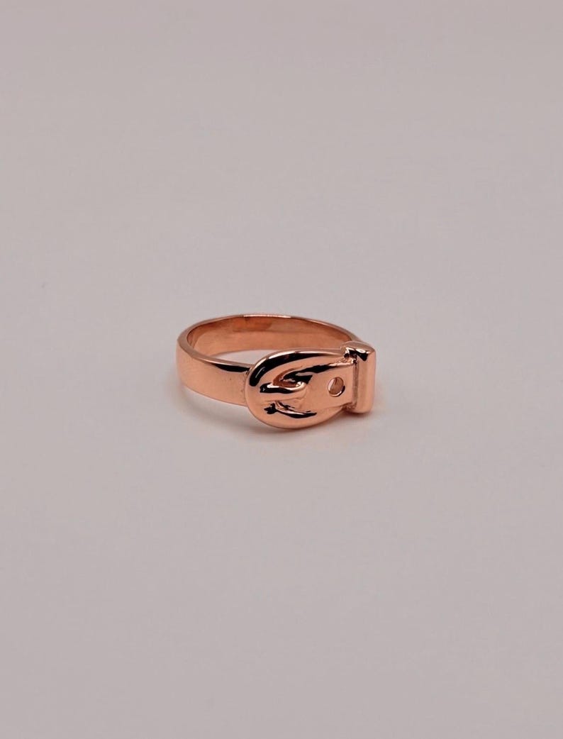 May include: A rose gold-toned metal ring designed to resemble a miniature belt. The band features a central buckle element with a prong and a small hole, mimicking the appearance of a functional belt wrapped around the finger in a smooth finish.
