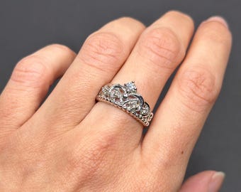 May include: A silver-colored ring in the shape of a crown, adorned with small clear gemstones. The ring has an openwork design with a larger gemstone at the top center. The ring is on a person's finger against a dark gray background.
