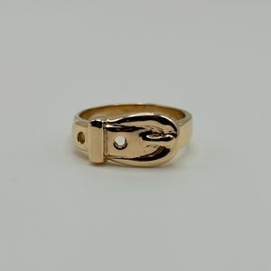 A gold-colored ring in the shape of a belt buckle. The ring has a detailed buckle design with a small hole on the side. The ring is made of 14K gold.