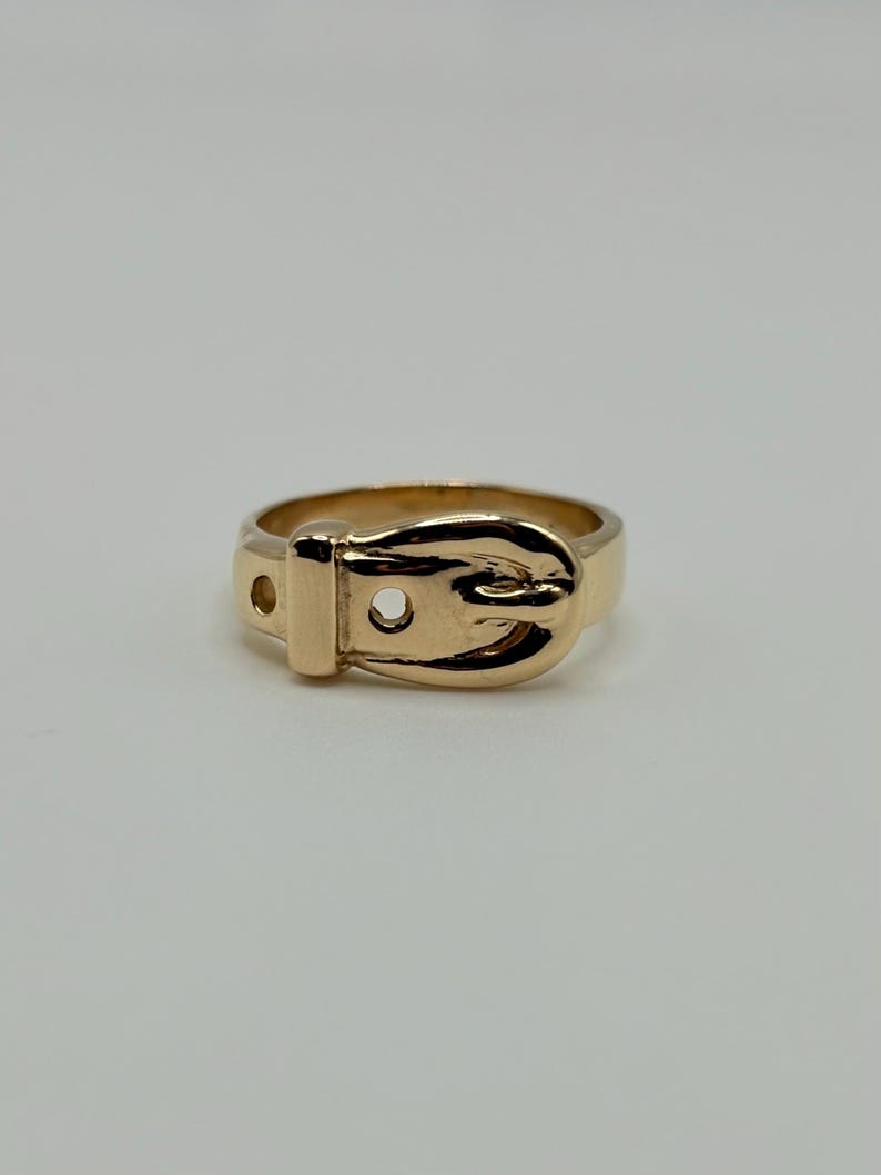 A gold-colored ring in the shape of a belt buckle. The ring has a detailed buckle design with a small hole on the side. The ring is made of 14K gold.