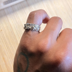 May include: A silver-toned ring with a textured, uneven surface featuring multiple small, raised, and irregular metallic shapes. The band has a wide, chunky profile with a reflective finish that catches the light across its complex, organic, and bumpy exterior design.