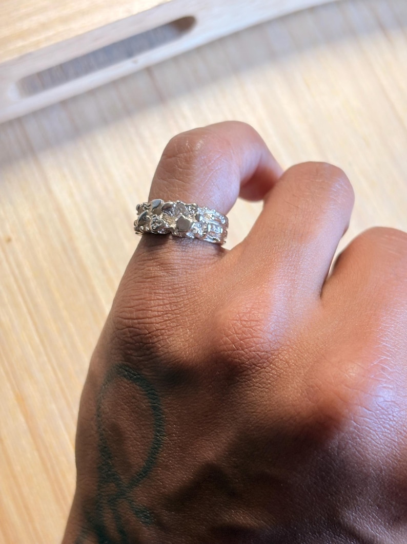 May include: A silver-toned ring with a textured, uneven surface featuring multiple small, raised, and irregular metallic shapes. The band has a wide, chunky profile with a reflective finish that catches the light across its complex, organic, and bumpy exterior design.
