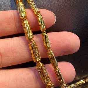 May include: Close-up of a gold necklace with a unique design. The necklace features rectangular links with intricate patterns. The gold chain is held by a person's hand, showcasing the details and craftsmanship of the jewelry.
