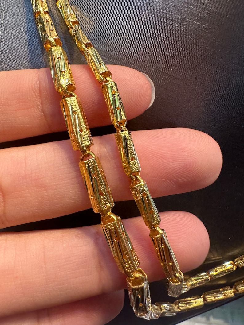 May include: Close-up of a gold necklace with a unique design. The necklace features rectangular links with intricate patterns. The gold chain is held by a person's hand, showcasing the details and craftsmanship of the jewelry.