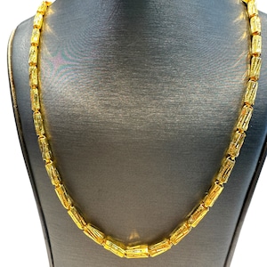May include: A gold-colored necklace with a repeating cylindrical design. The necklace is displayed on a gray mannequin. The chain links are rectangular with etched details, creating a textured appearance. The necklace is a bright, shiny gold.