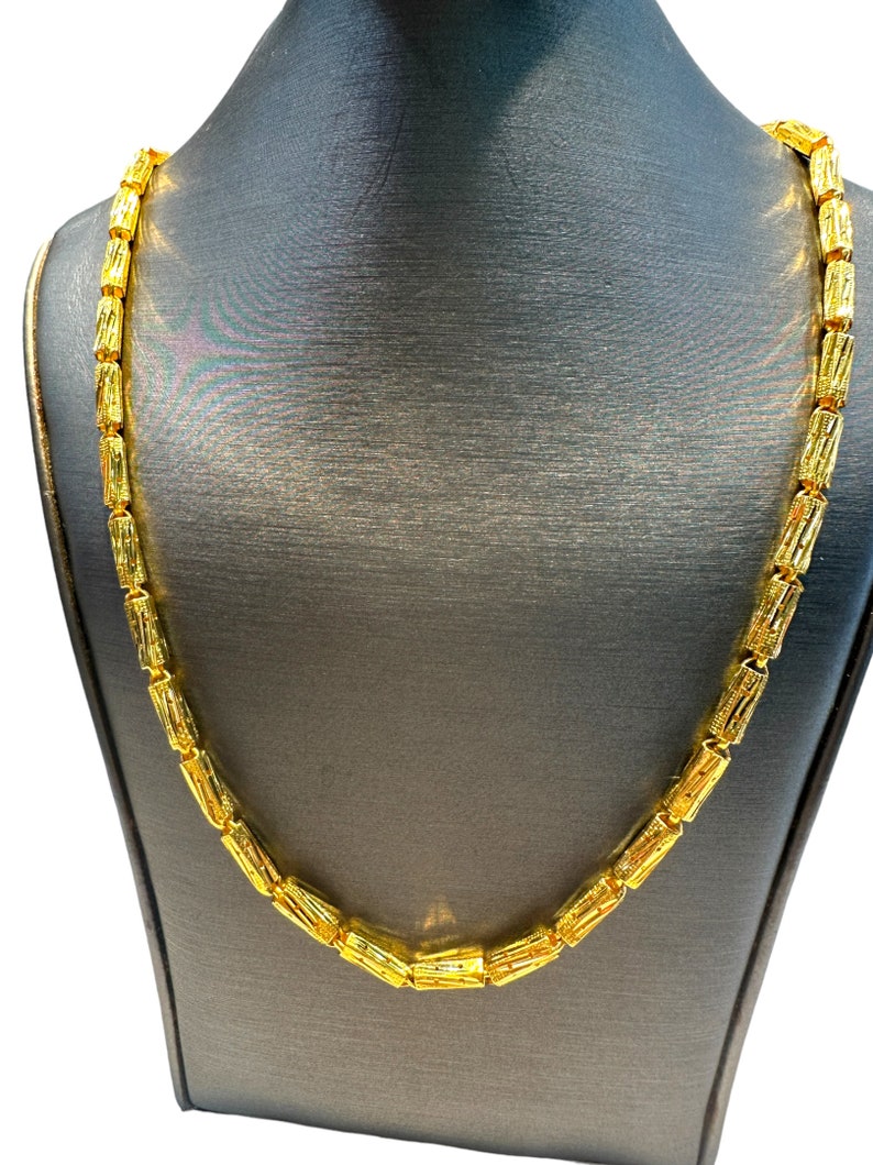 May include: A gold-colored necklace with a repeating cylindrical design. The necklace is displayed on a gray mannequin. The chain links are rectangular with etched details, creating a textured appearance. The necklace is a bright, shiny gold.