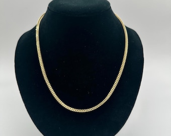 May include: A gold-colored chain necklace displayed on a black mannequin. The chain has a tightly linked design, creating a textured appearance. The necklace is draped in a curved shape, showcasing its form and style. The background is a plain white.