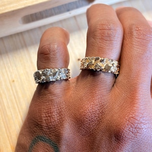 May include: Two gold-toned rings with a textured, nugget-like surface finish. Each ring features an irregular, organic design with raised metallic segments that catch the light, creating a rugged appearance across the entire band of the jewellery.