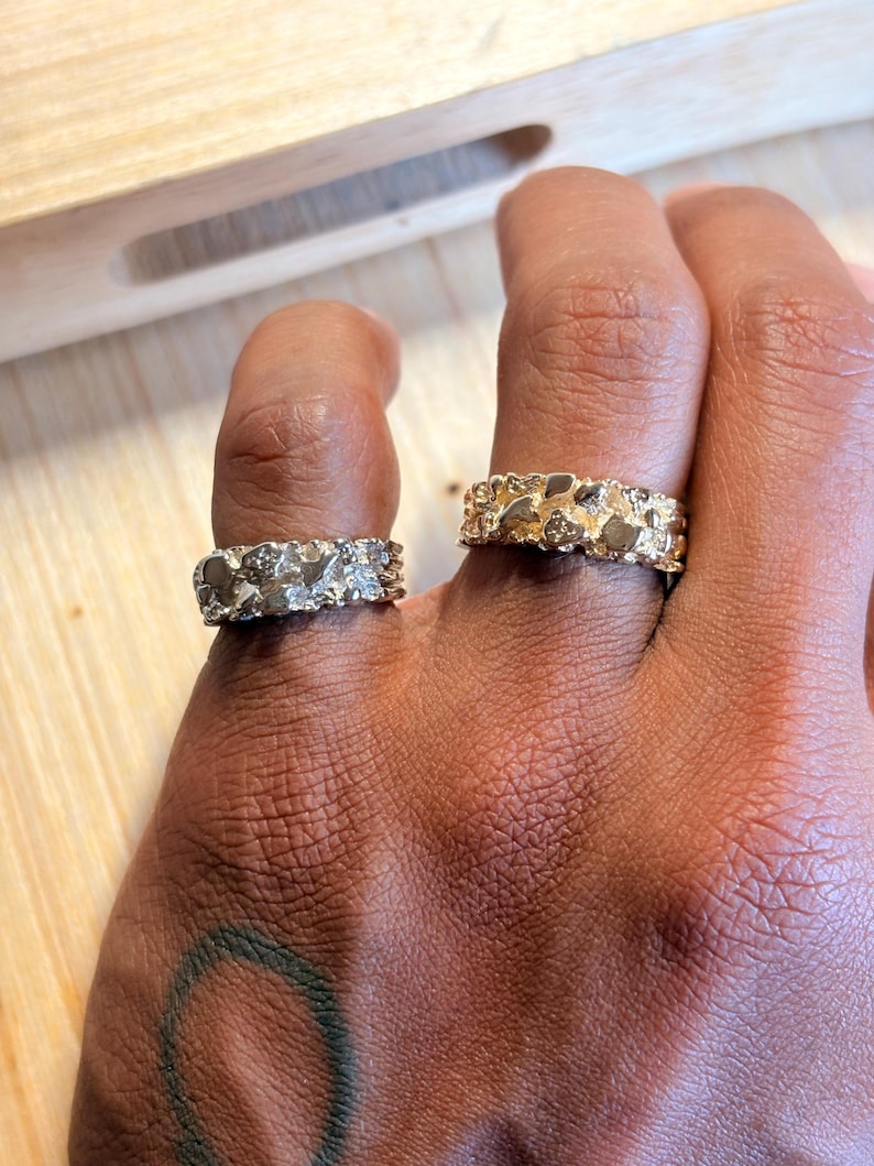 May include: Two gold-toned rings with a textured, nugget-like surface finish. Each ring features an irregular, organic design with raised metallic segments that catch the light, creating a rugged appearance across the entire band of the jewellery.