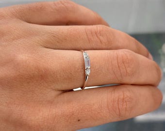 May include: A silver-toned ring featuring a central baguette-cut stone flanked by two smaller round-cut stones. The band is thin and polished, showcasing a minimalist design with three clear, faceted stones set horizontally along the top.