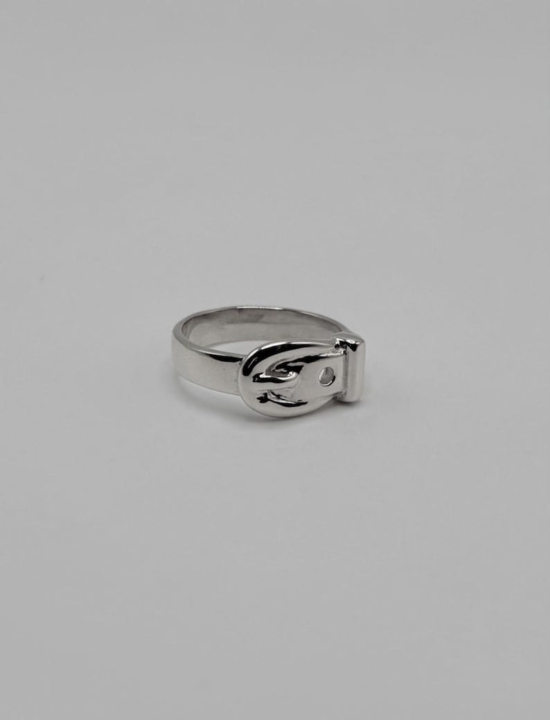 May include: A polished silver-toned ring featuring a design that mimics a belt, complete with a buckle-shaped focal point on the front. The band is smooth and uniform, showcasing a reflective metallic finish throughout the entire piece.