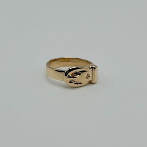 A gold-colored ring with a buckle design. The ring features a detailed buckle shape, with a rectangular base and a curved top. The ring is set against a plain white background.