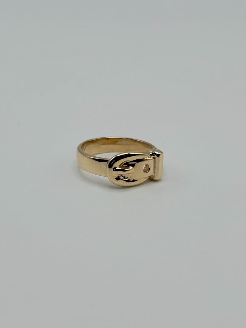 A gold-colored ring with a buckle design. The ring features a detailed buckle shape, with a rectangular base and a curved top. The ring is set against a plain white background.