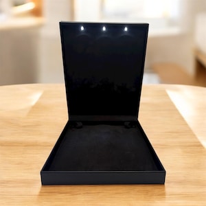 May include: Open black jewelry box with three small lights inside the lid. The box is open, revealing a black velvet interior. The box is sitting on a light brown wooden surface.