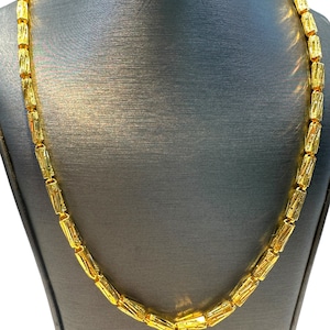 May include: A gold-colored necklace with a repeating cylindrical bead design. The necklace is displayed on a gray mannequin, showcasing its length and style. The beads have a textured surface, reflecting light.
