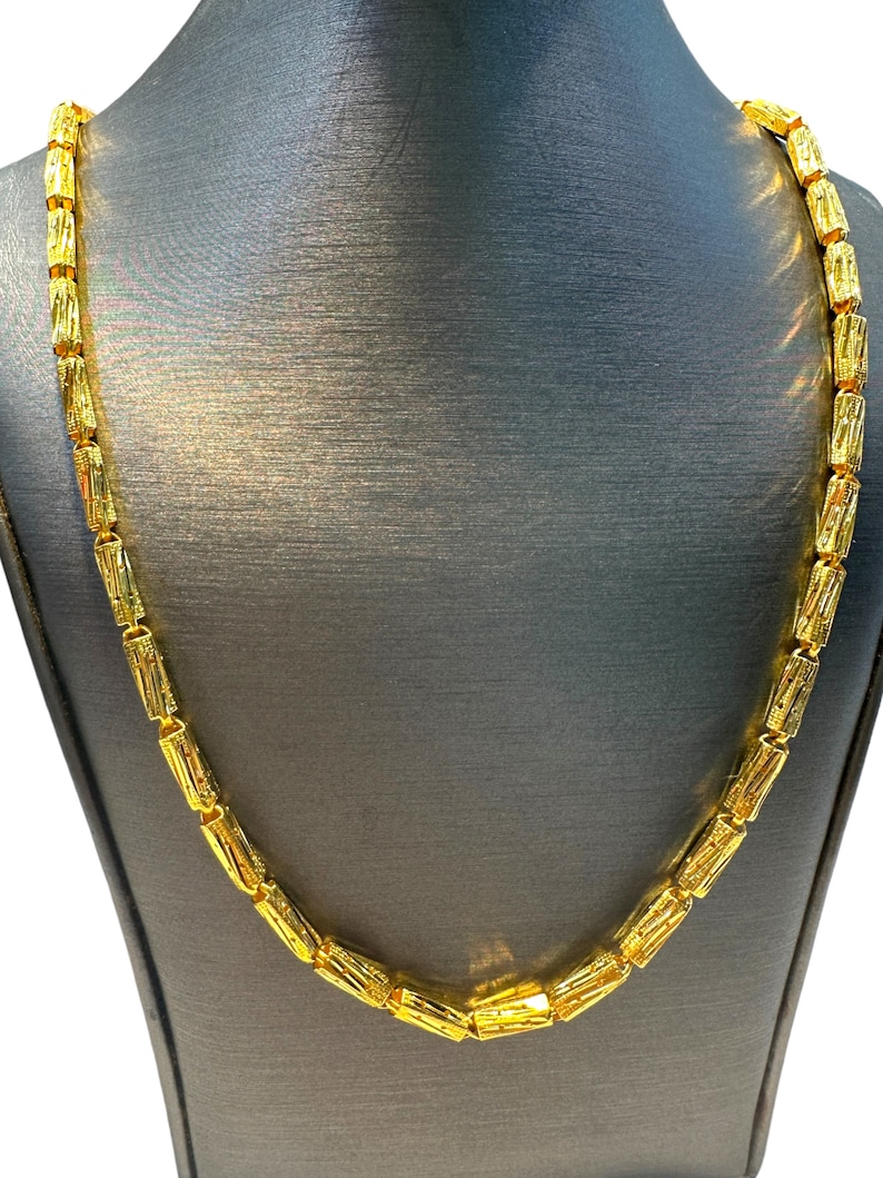 May include: A gold-colored necklace with a repeating cylindrical bead design. The necklace is displayed on a gray mannequin, showcasing its length and style. The beads have a textured surface, reflecting light.