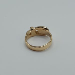 A gold-colored ring with a buckle design. The ring has a smooth, polished band and a detailed buckle element. The ring is set against a plain, light background.