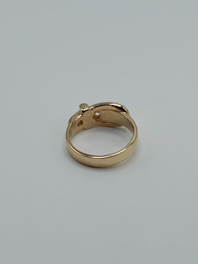 A gold-colored ring with a buckle design. The ring has a smooth, polished band and a detailed buckle element. The ring is set against a plain, light background.