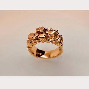 May include: A gold ring featuring a band with a highly textured, irregular surface composed of multiple raised, rounded, and molten-looking metallic shapes. The metal has a polished, reflective finish that catches the light across its uneven, organic form.